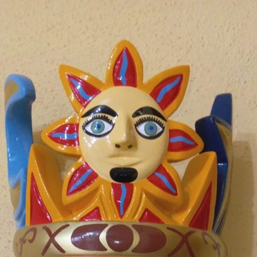 Acercamiento a la corona de la máscara de El Tiempo: sol amarillo con cara y rayos rojos y azules sobre una banda dorada