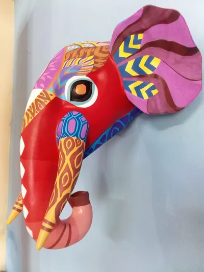 Máscara de elefante de perfil, roja con oreja rosa, trompa enroscada y patrones de colores