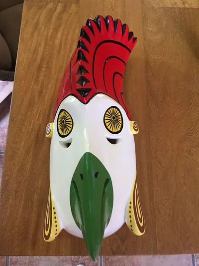 Máscara del gallo rojo, de rostro blanco con cresta roja, ojos amarillos de rayos negros y pico verde, sobre una mesa de madera