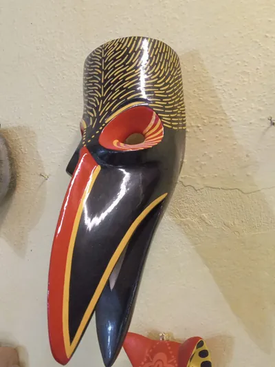 Máscara de ave negra con pico largo bordeado de rojo y plumas pintadas con trazos amarillos, vista de lado
