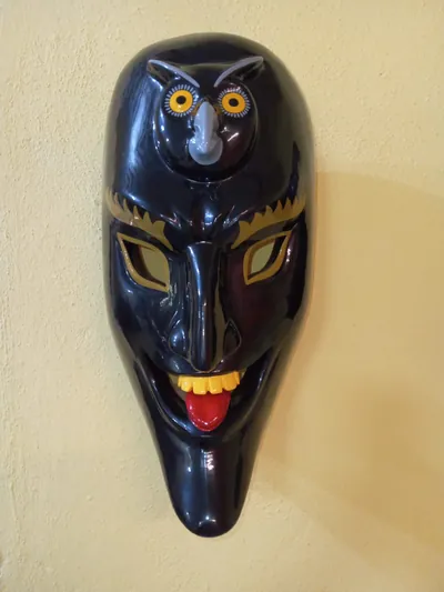Máscara negra de rostro alargado con lengua roja de fuera y un búho de ojos amarillos tallado en la frente