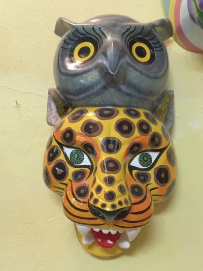 Máscara de jaguar amarillo con manchas oscuras, colmillos y lengua roja, coronada por un búho gris de ojos amarillos