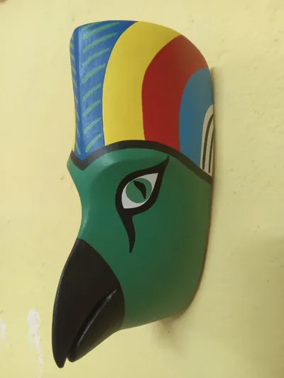 Máscara de guacamaya verde de perfil con pico negro y copete de franjas azul, amarilla y roja