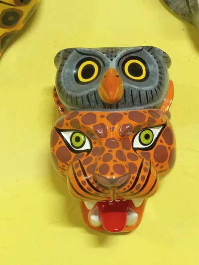 Máscara de felino anaranjado con manchas café y lengua roja, coronada por un búho gris de pico anaranjado