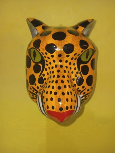 Máscara de jaguar anaranjado con manchas negras, ojos verdes de pupila rasgada y nariz roja, de frente sobre pared amarilla