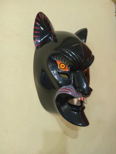 Máscara de nahual negro con cara de felino, ojo anaranjado, orejas con rayas rojas y colmillos, vista de lado