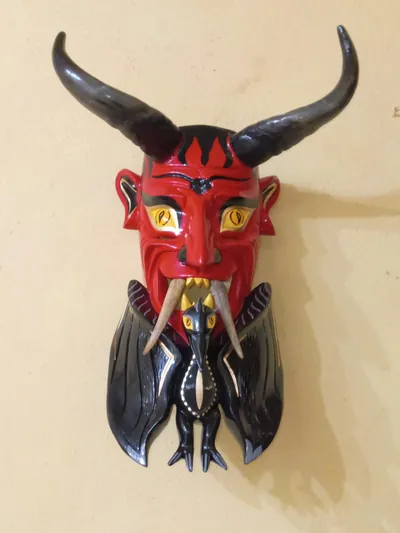 Máscara de diablo rojo con cuernos negros, ojos amarillos y una criatura negra alada bajo la barbilla