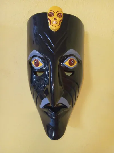 Máscara negra de rostro alargado con arrugas, bigote gris y una pequeña calavera amarilla en la frente