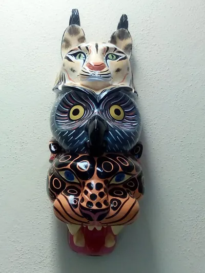 Máscara de madera con un lince, un búho y un jaguar de rosetas tallados uno encima de otro, sobre pared clara