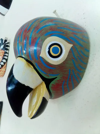 Máscara de guacamaya roja y verde con trazos azules y pico ganchudo negro y crema, vista de lado