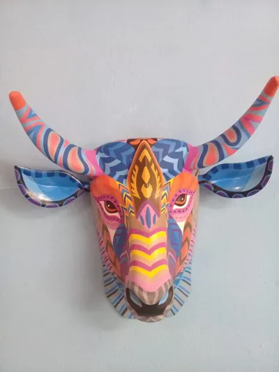 Máscara de toro de frente con cuernos y orejas pintados de grecas azules, rosas y amarillas sobre pared azul