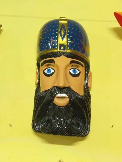 Máscara de hombre con barba negra y gorro azul con estrellas, sol y luna dorados sobre pared amarilla