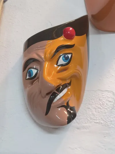 Máscara de hombre de rostro partido en piel rosada y anaranjada, con bigote negro y bola roja en la frente