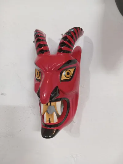 Máscara de diablo rojo con cuernos rayados en negro, ojos amarillos y boca abierta con colmillos, en pared blanca