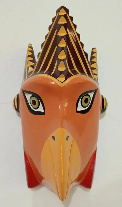 Máscara de gallina anaranjada con cresta color vino escalonada, pico amarillo y barbas rojas, de frente en pared blanca