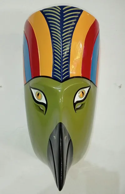 Máscara de guacamaya verde con pico negro y copete de bandas rojas, amarillas y azules, de frente en pared blanca