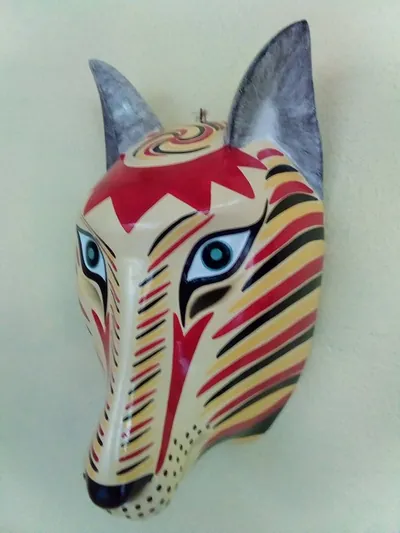 Máscara de lince crema con rayas rojas y negras, zigzag rojo en la frente y ojos azules, vista de lado en pared crema
