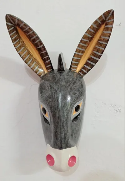 Máscara de burro gris de frente, con orejas largas rayadas y hocico blanco con fosas nasales rosas, en pared blanca