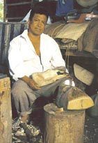 Don Herminio Candelario Dolores, con camisa blanca, sentado en su taller trabaja una pieza de madera apoyada en un tronco